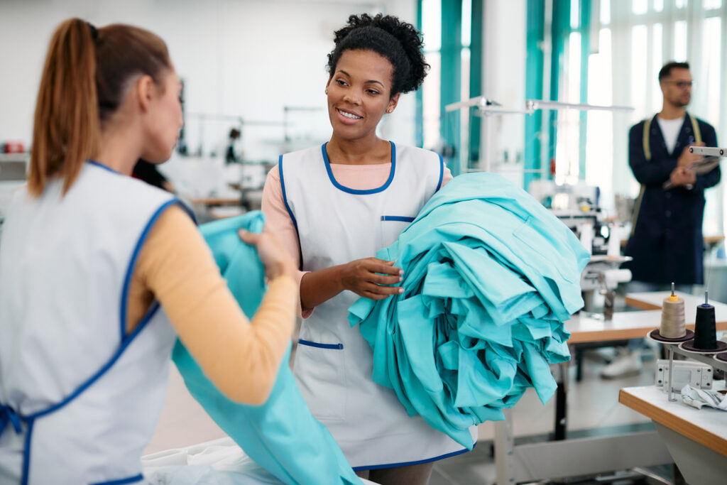 Young black dressmaker communicating with female coworker while working in textile factory